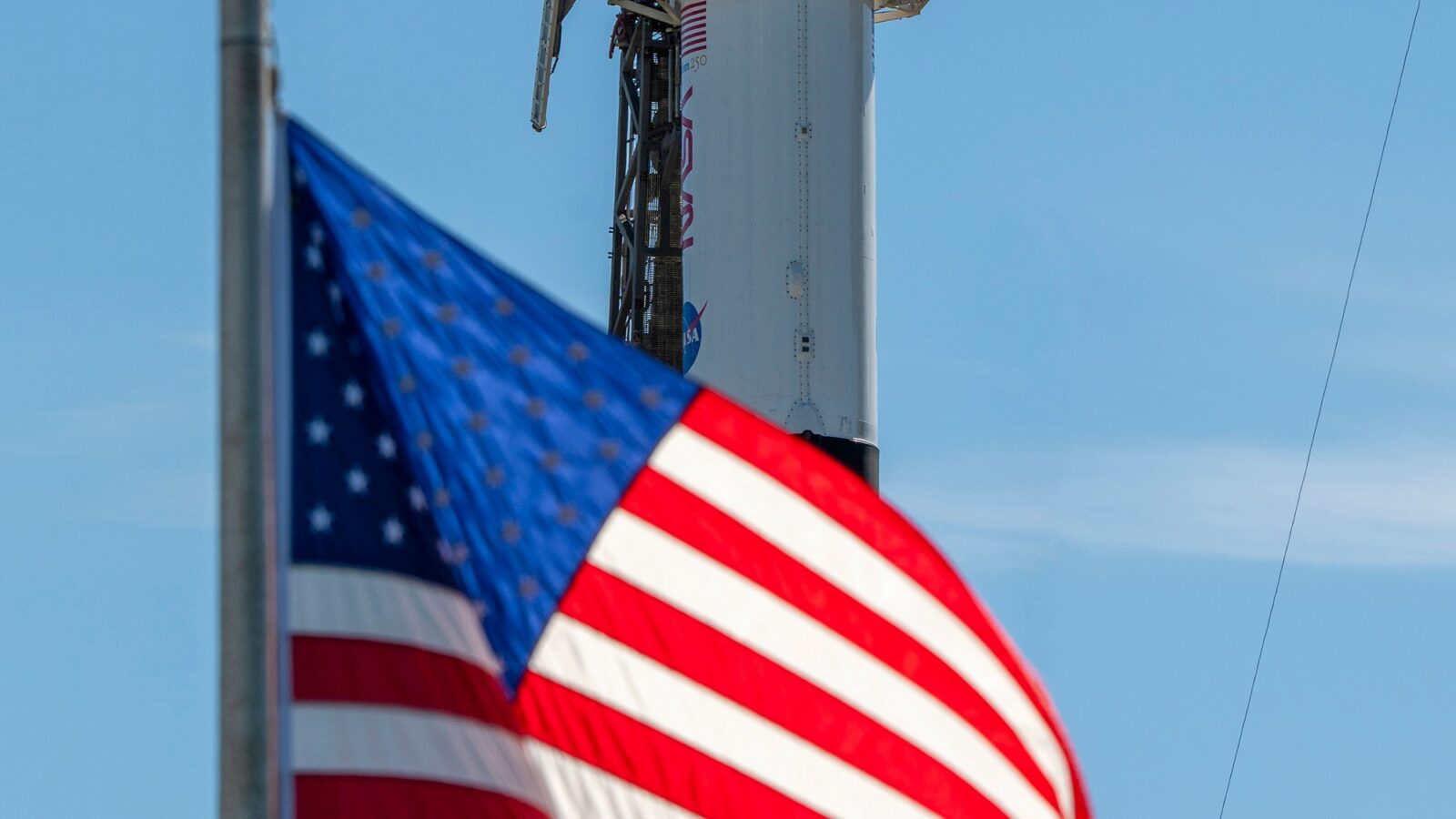 A close-up view of a SpaceX Crew Dragon spacecraft mounted atop a Falcon 9 rocket on the launch pad. The capsule and trunk are connected to ground support hardware, with NASA and mission insignia visible on the rocket’s exterior against a bright blue sky. in the foreground in the American flag.
