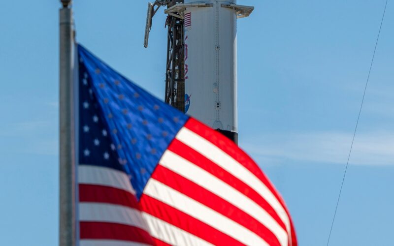 A close-up view of a SpaceX Crew Dragon spacecraft mounted atop a Falcon 9 rocket on the launch pad. The capsule and trunk are connected to ground support hardware, with NASA and mission insignia visible on the rocket’s exterior against a bright blue sky. in the foreground in the American flag.