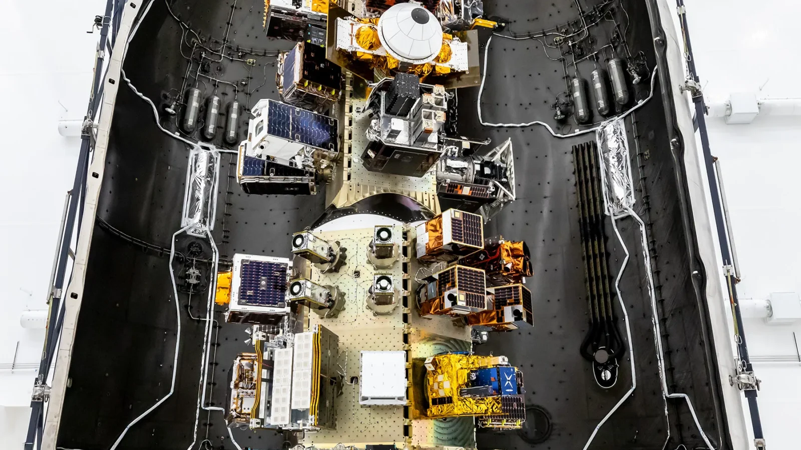 Top‑down view of a SpaceX payload fairing interior showing mounted satellites and hardware arranged along the center structure, surrounded by the curved black fairing walls.
