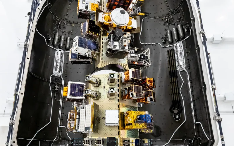 Top‑down view of a SpaceX payload fairing interior showing mounted satellites and hardware arranged along the center structure, surrounded by the curved black fairing walls.