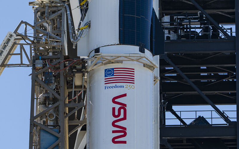 A close-up view of a SpaceX Crew Dragon spacecraft mounted atop a Falcon 9 rocket on the launch pad. The capsule and trunk are connected to ground support hardware, with NASA and mission insignia visible on the rocket’s exterior against a bright blue sky.